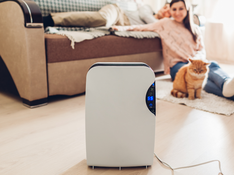 A humidifier or dehumidifier placed next to a cozy bed in a bedroom, with a visible humidity reading on a nearby digital thermometer.